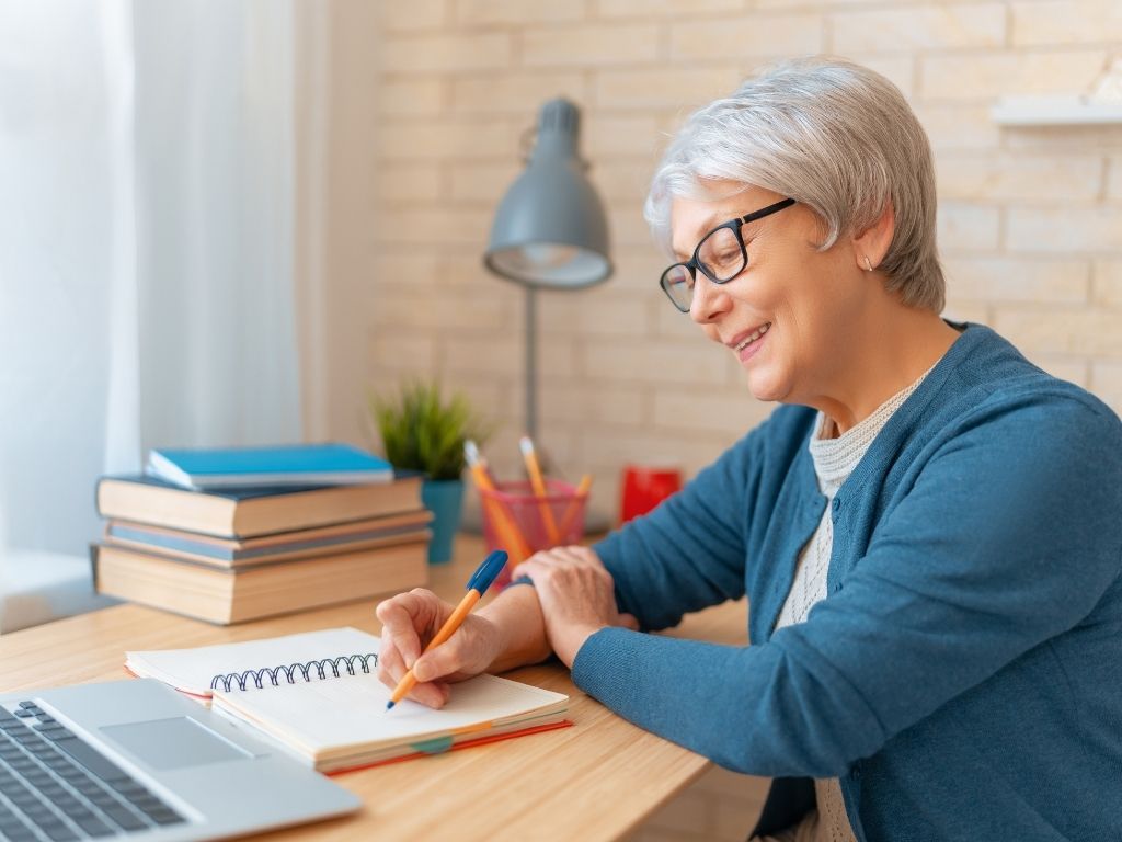 An older woman smiling and writing in a notebook in front of a computer.